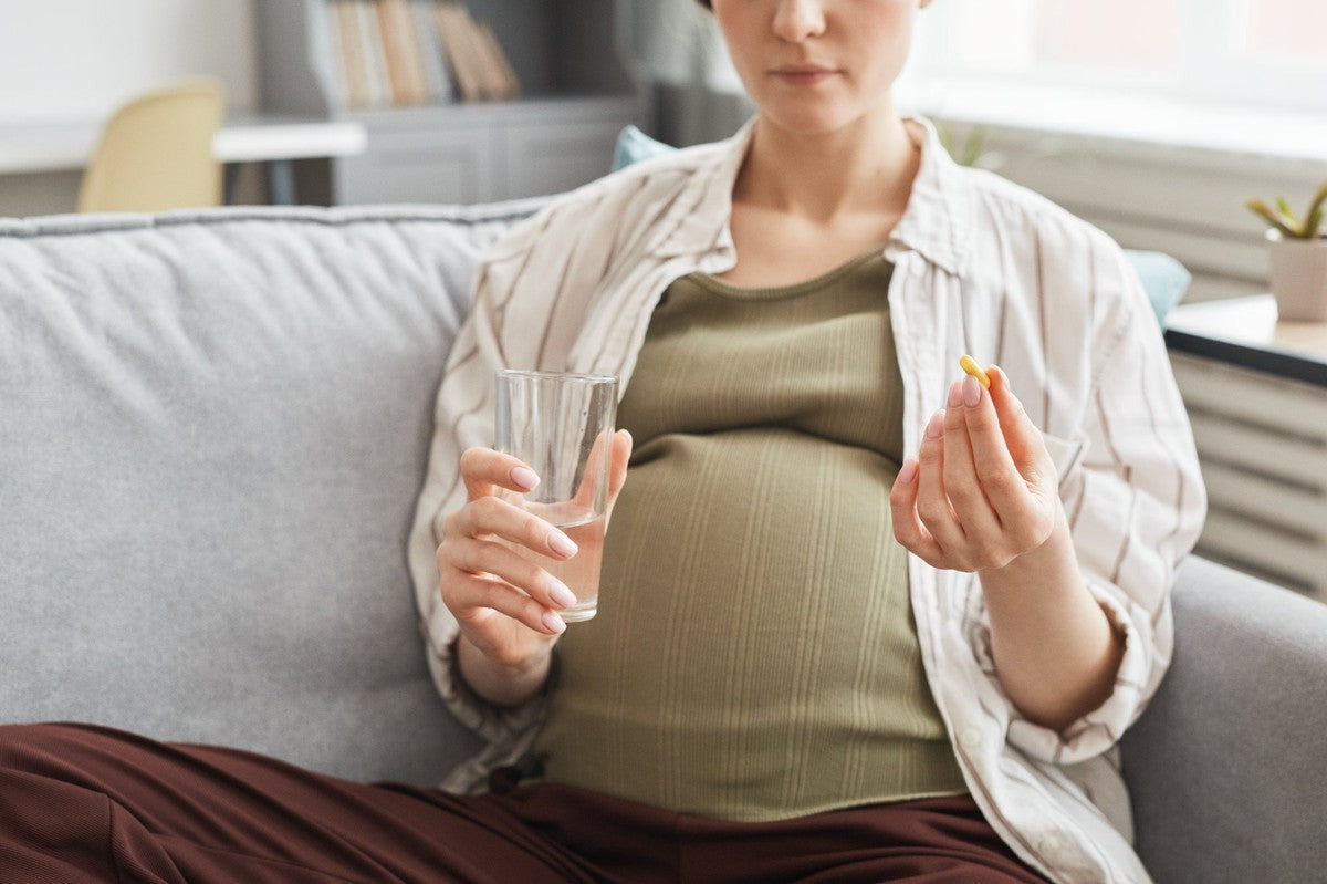 A pregnant woman on a sofa, holding a glass of water and an iron capsule, considering her daily supplement routine