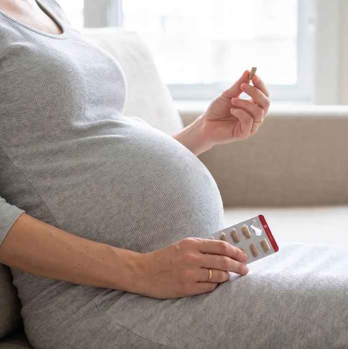 Pregnant woman on a sofa hesitating before taking a prenatal iron supplement capsule from a blister pack